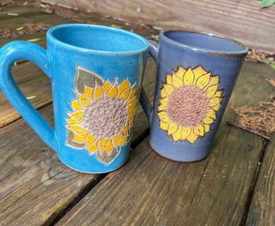 Handmade pottery mugs with blue glazes and carved out sunflowers that are painted in yellow.