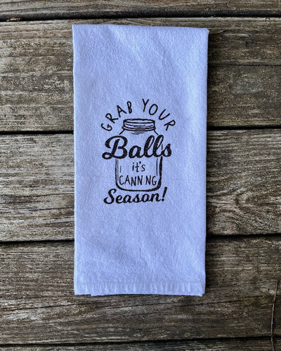 A white dish towel with a black glitter vinyl design that says "Grab your balls it's canning season" with a balls canning jar.  A funny and useful kitchen gifts for those who like home canning.