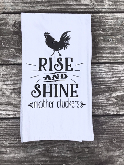 A white cotton kitchen towel with funny farmhouse design of a rooster and quote "rise and shine mother cluckers" in black vinyl.  A funny and useful kitchen gift.